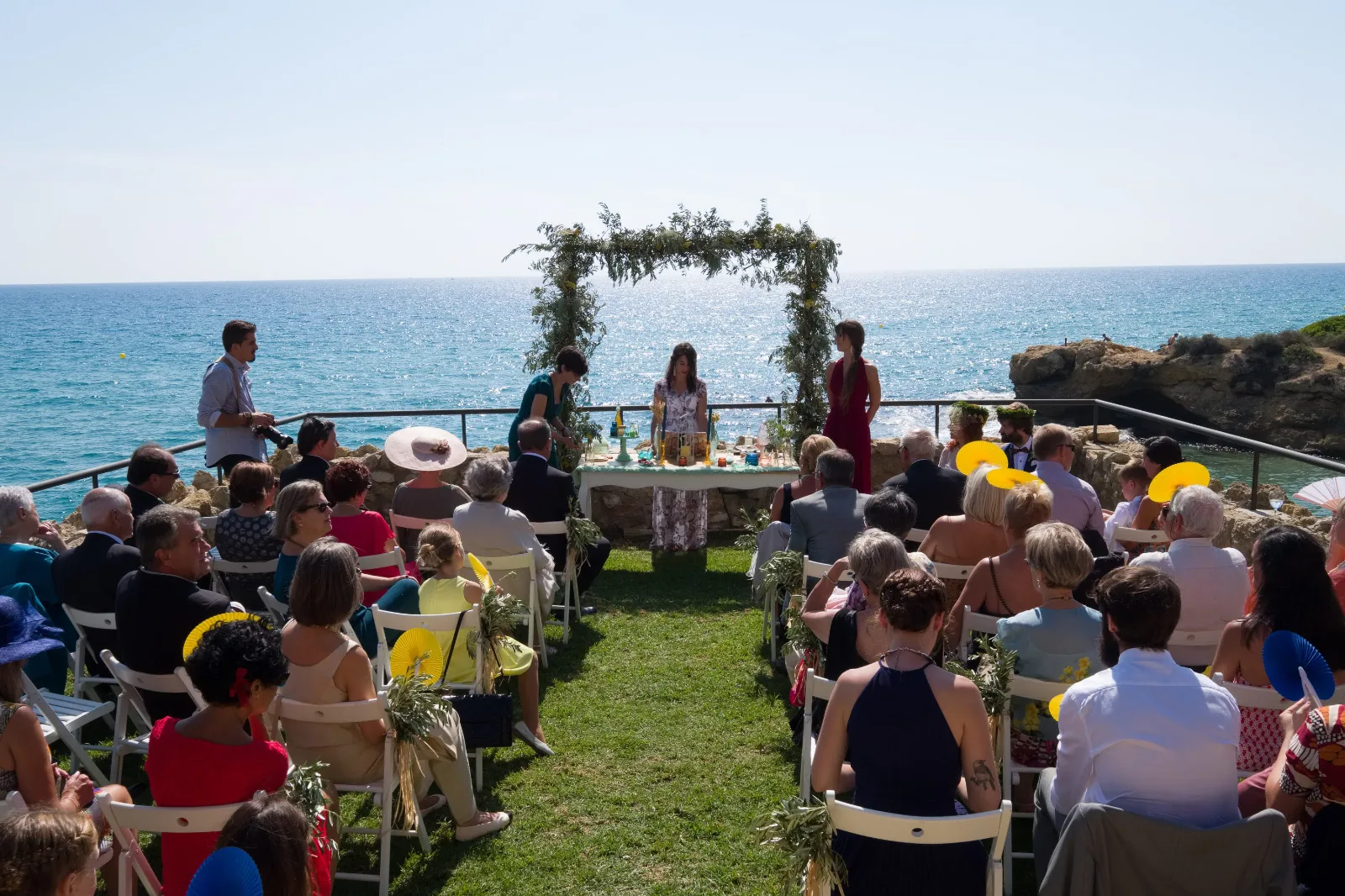 Cerimònia de casament a l’aire lliure davant del mar, amb un arc de vegetació i convidats asseguts